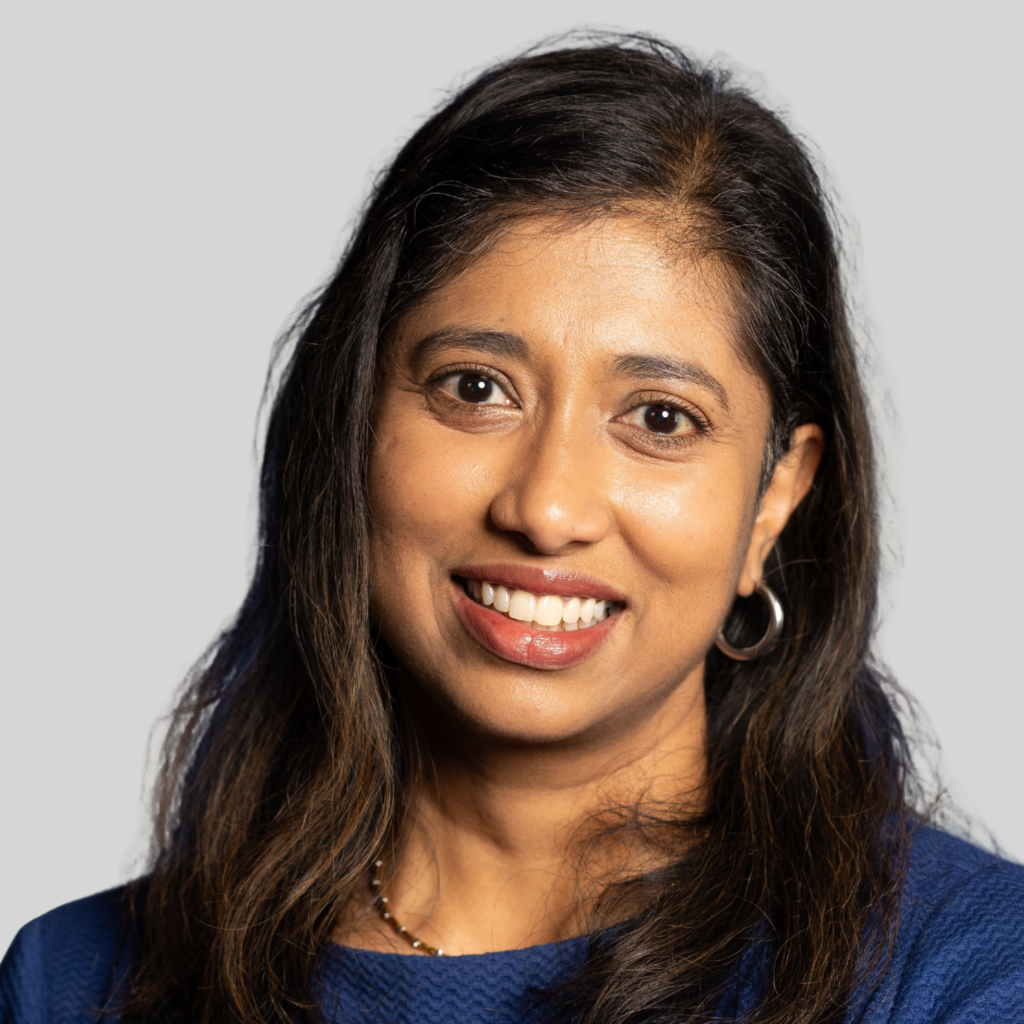 A woman with long dark hair, wearing a blue top and hoop earrings, smiles at the camera against a light gray background, ready to join the RSNA event’s Lunch & Learn session.