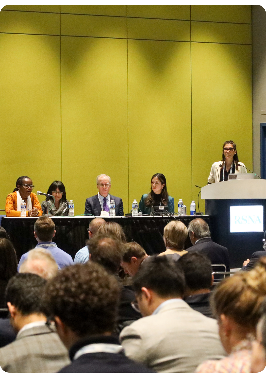 Five panelists and a moderator are seated at a table onstage, facing an audience in a conference room with yellow-green walls. A sign on the podium reads “RSNA.”.